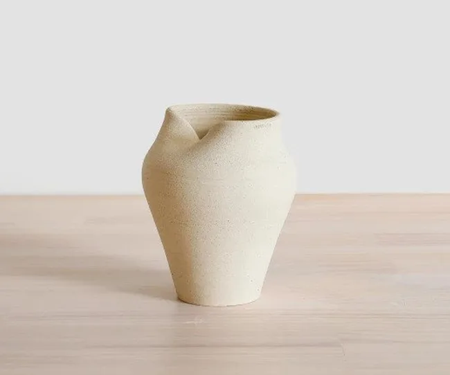 Cream-colored vase with a unique twisted top, placed on a light wooden surface against a plain background.