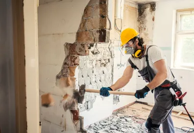 Construction worker in safety gear using a sledgehammer to demolish a wall in a partially stripped room.