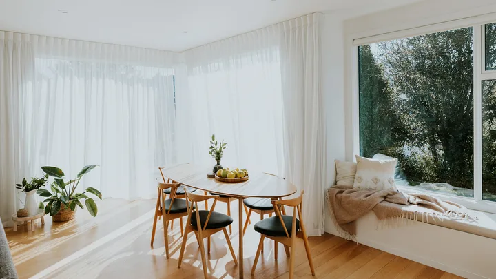 Bright dining room with wooden table, chairs, plants, and large windows with sheer curtains allowing natural light.