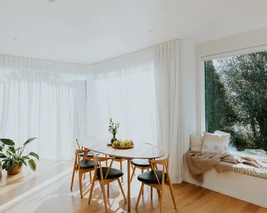 Bright dining room with wooden table, chairs, plants, and large windows with sheer curtains allowing natural light.
