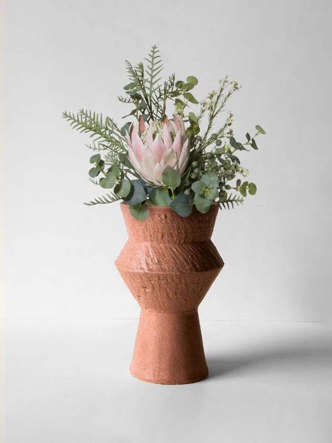 A geometric ceramic vase holding a pink protea and greenery, set against a plain background.