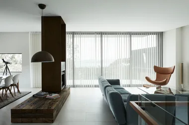 Modern living room with blue sofa, wooden table, brown chair, pendant light, and large windows with sheer curtains.