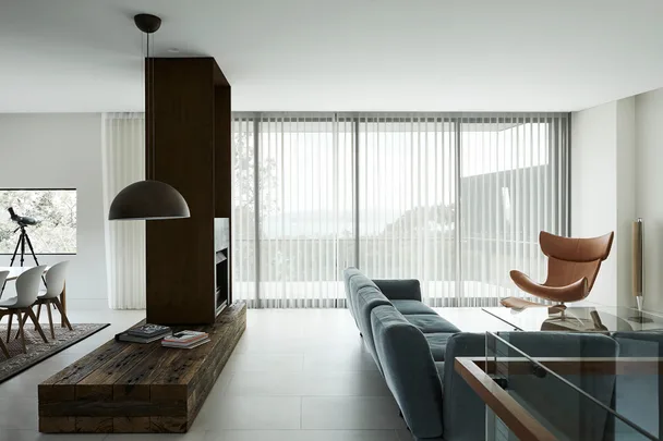 Modern living room with blue sofa, wooden table, brown chair, pendant light, and large windows with sheer curtains.