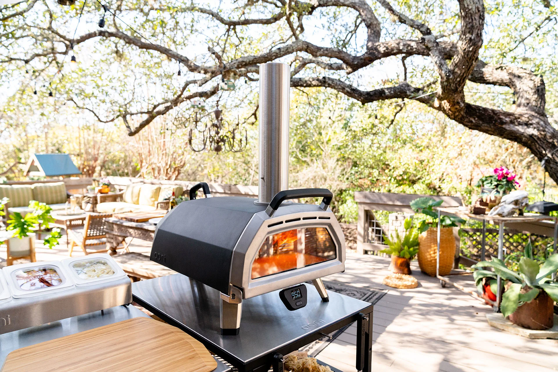 Outdoor scene with a black tabletop pizza oven, wooden table, and lush greenery.
