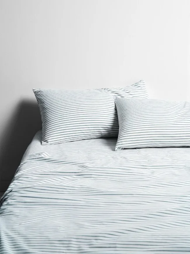 Striped bed with white and gray bedding, two pillows against a plain white wall.