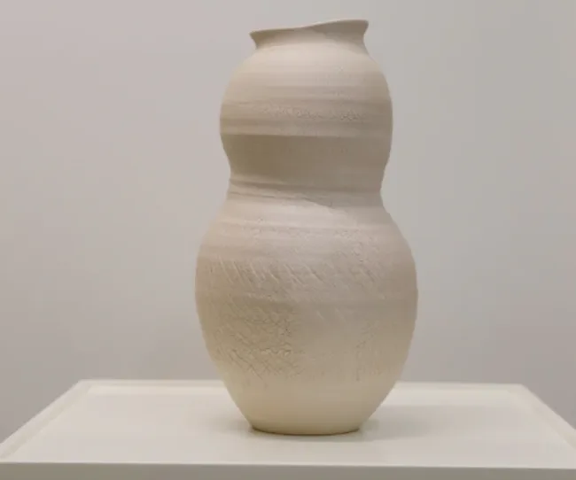 A cream-colored, textured ceramic vase with a unique, double-gourd shape on a white pedestal.