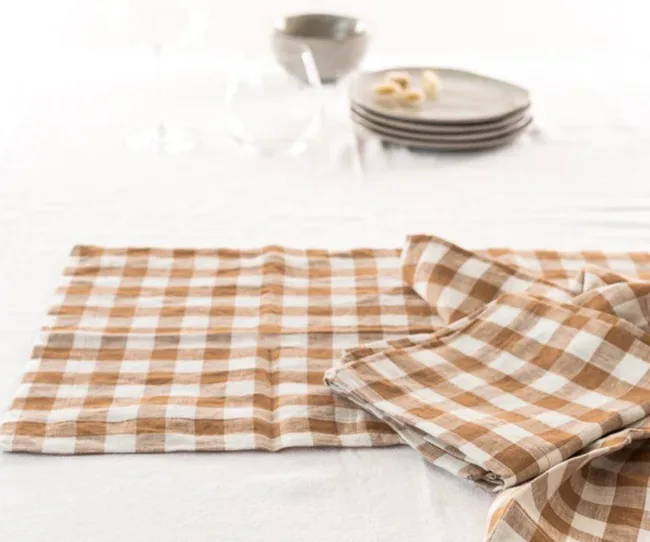 Brown and white checkered tablecloth folded on a white tablecloth with stacked plates in the background.