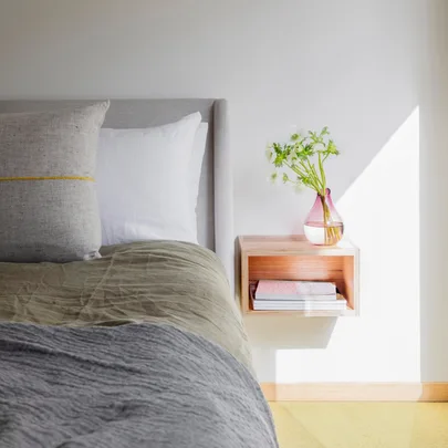 Cozy bedroom scene with a bed, gray and white pillows, green bedspread, wall shelf, pink vase with flowers, and books.