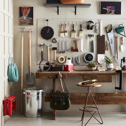 Workshop with tools organized on a pegboard, wooden workbench, hose, and plants; art on the walls.