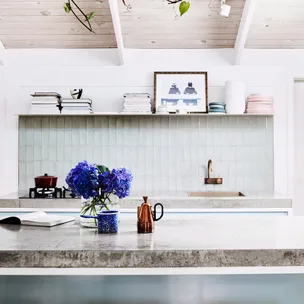Modern kitchen with gray countertops, blue hydrangeas, and a copper kettle. Shelves with books and dishes are in the background.