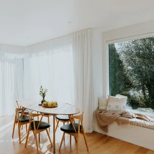 Bright dining room with wooden table and chairs, fruit bowl centerpiece; large window with tree view and bench seat.