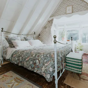 Cozy attic bedroom with floral bedding and wallpaper, iron bed frame, wooden floor, and window with potted plants.
