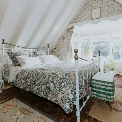 Cozy attic bedroom with floral bedding and wallpaper, iron bed frame, wooden floor, and window with potted plants.