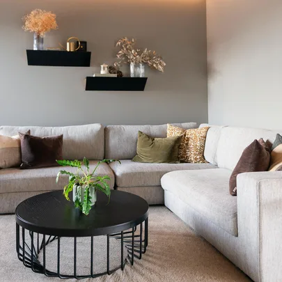 Gray sectional sofa with assorted pillows, black round coffee table with plant, and wall shelves in a cozy living room.