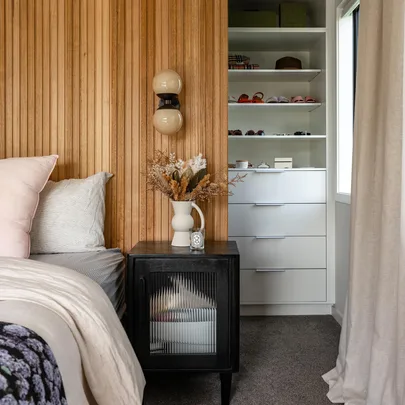 Cozy bedroom corner with a wooden accent wall, modern nightstand, decorative vase, and open shelves with accessories.