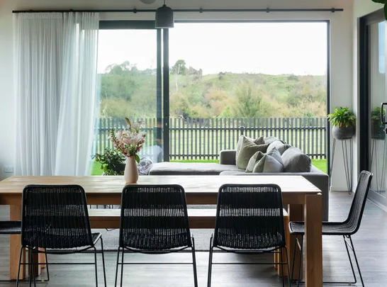 Modern living room with a wooden dining table, black chairs, and a large glass window overlooking a garden.