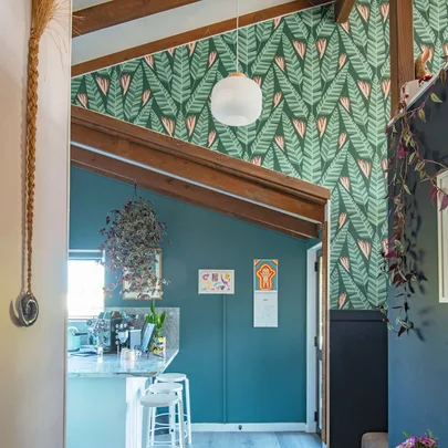 Interior with tropical leaf wallpaper, wooden beams, blue walls, hanging plant, kitchen counter, and colorful wall art.