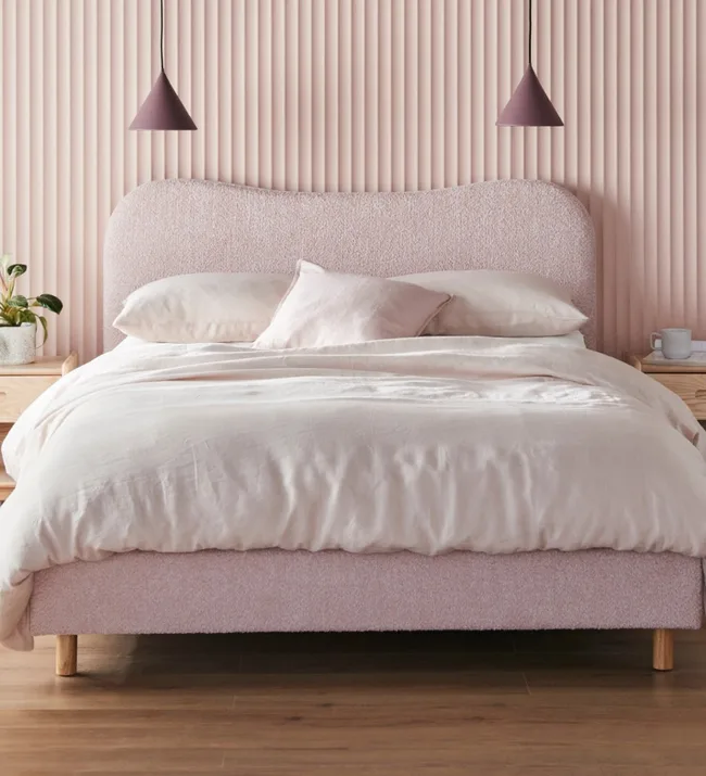 Cozy pink bed with soft linens, wooden bedside table, pendant lights, and potted plant on hardwood floor.