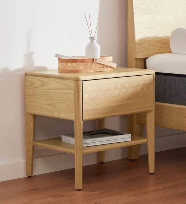 Wooden bedside table with a drawer, a lower shelf holding books, and a vase with sticks on top.