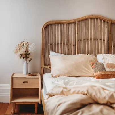 Cozy bedroom with rattan headboard, beige bedding, a decorative pillow, and a nightstand with dried flowers in a vase.