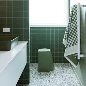 Green-tiled bathroom with a geometric stool, checkered towel, and a large basin. Bright window lighting the space.