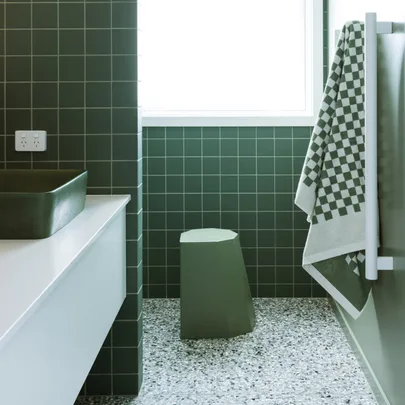Green-tiled bathroom with a geometric stool, checkered towel, and a large basin. Bright window lighting the space.