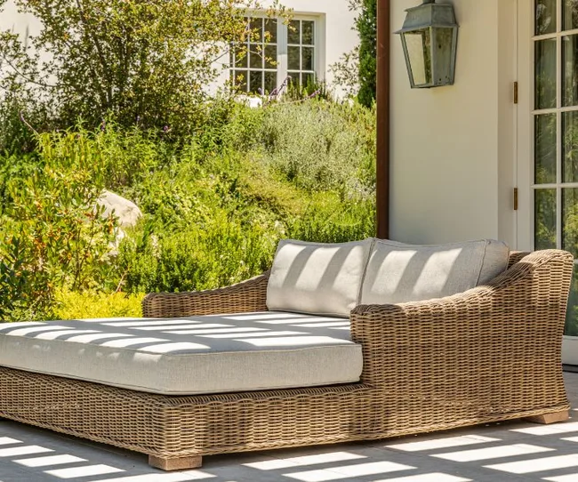 Wicker outdoor sofa with cushions on a patio, surrounded by lush greenery and a white house wall in the background.