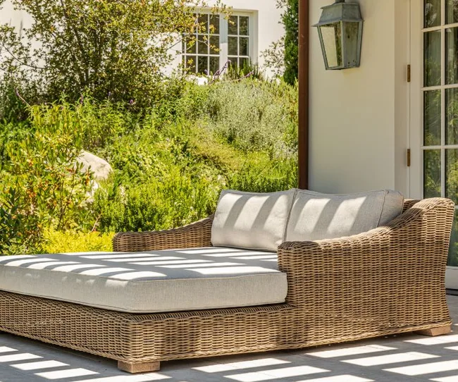 Wicker outdoor sofa with cushions on a patio, surrounded by lush greenery and a white house wall in the background.