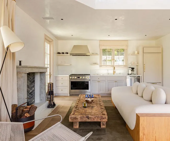 Cozy living room with a rustic wooden coffee table, modern couch, fireplace, and an open kitchen area in soft tones.