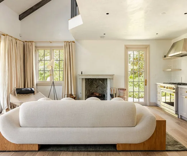 Spacious living room with a white couch, fireplace, and modern kitchen appliances, natural light from large windows.