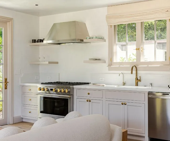 Modern kitchen with white cabinets, stainless steel appliances, wood shelves, and large window above the sink.