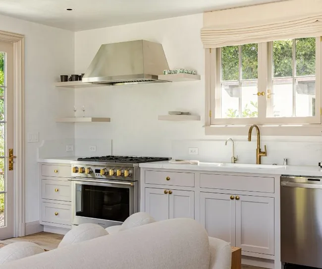 Modern kitchen with white cabinets, stainless steel appliances, wood shelves, and large window above the sink.
