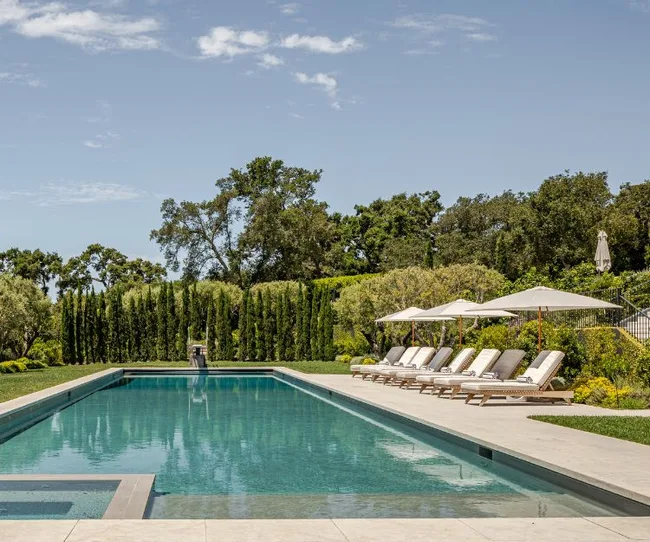 Luxurious outdoor swimming pool with lounge chairs, umbrellas, and surrounded by lush greenery under a clear sky.