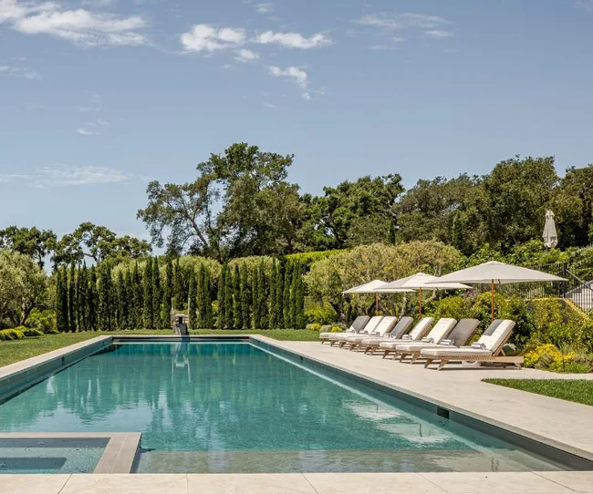 Luxurious outdoor swimming pool with lounge chairs, umbrellas, and surrounded by lush greenery under a clear sky.