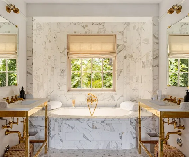 Luxurious marble bathroom with gold fixtures, large window showing greenery, and two sinks flanking a central bathtub.