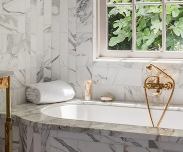 Luxurious marble bathroom with tub, gold fixtures, rolled white towels, and daylight through the window.