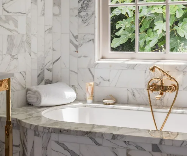 Luxurious marble bathroom with tub, gold fixtures, rolled white towels, and daylight through the window.