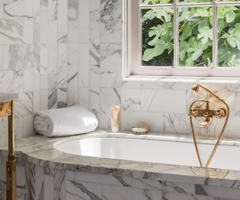 Luxurious marble bathroom with tub, gold fixtures, rolled white towels, and daylight through the window.