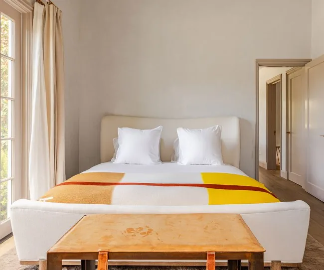 Cozy bedroom with a minimalist design, featuring a bed with white pillows and a colorful blanket, near a window.