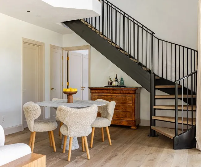 Dining area with a round table, shearling chairs, and a wooden chest under a black metal spiral staircase.