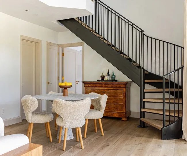 Dining area with a round table, shearling chairs, and a wooden chest under a black metal spiral staircase.