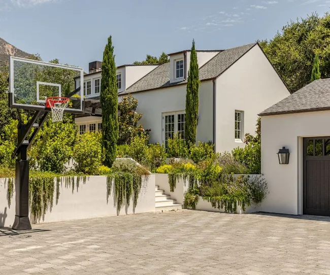 White house with tall trees, a basketball hoop, and a stone driveway under a clear sky.