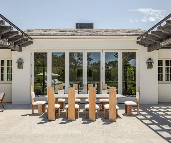 Outdoor dining area with wooden table and chairs, set on a stone patio in front of a glass-paneled house facade.