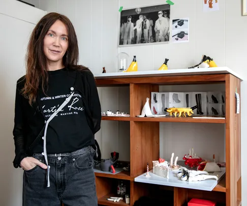 Woman in a black shirt stands next to a wooden shelf displaying artistic shoe miniatures and sculptures.