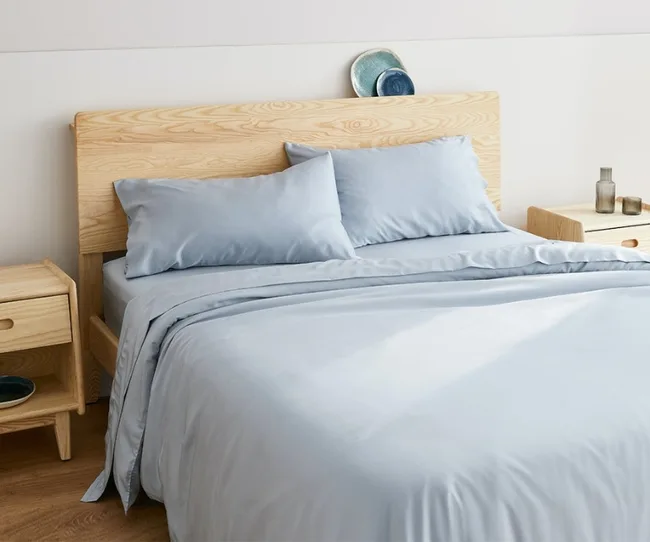 A bed with light blue sheets and pillows, a wooden headboard, and matching nightstands in a minimalist room.