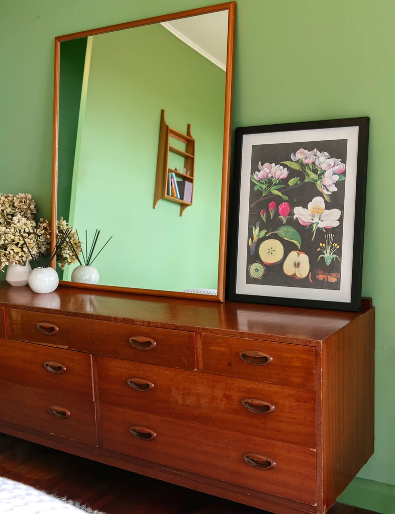 Large mirror above wooden dresser with floral artwork, vases, and green wall reflection in a cozy room setup.