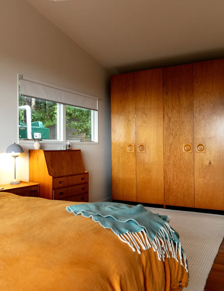 Cozy bedroom with wooden furniture, a large wardrobe, desk, bedside lamp, window, and an orange bed with a teal blanket.