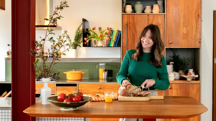 Woman in a green sweater slices bread in a bright, cozy kitchen with wooden cabinets and colorful decor.