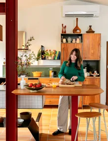 Woman in a green sweater slices bread in a bright, cozy kitchen with wooden cabinets and colorful decor.