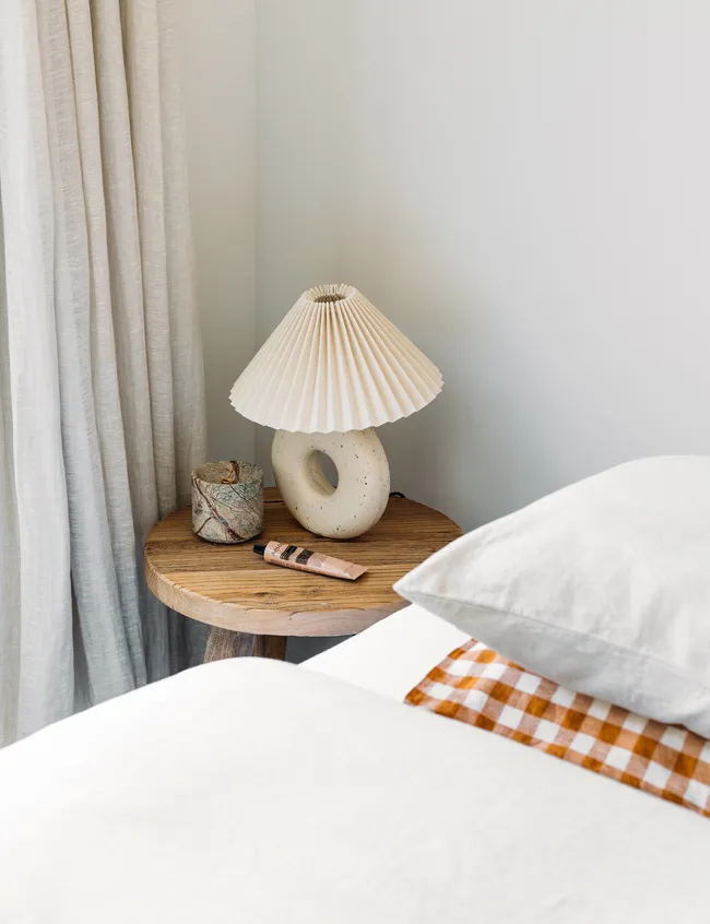 Bedside table with a modern lamp, a small container, and a tube of cream. White bedding and checkered pillow visible.